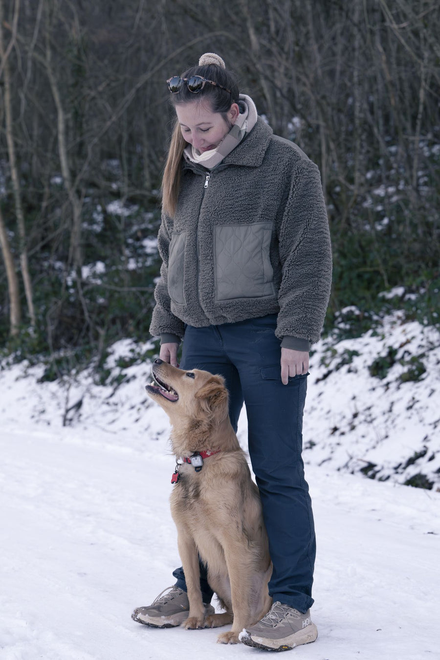 Une éducatrice debout dans la neige, un chien roux assis à ses pieds la regarde